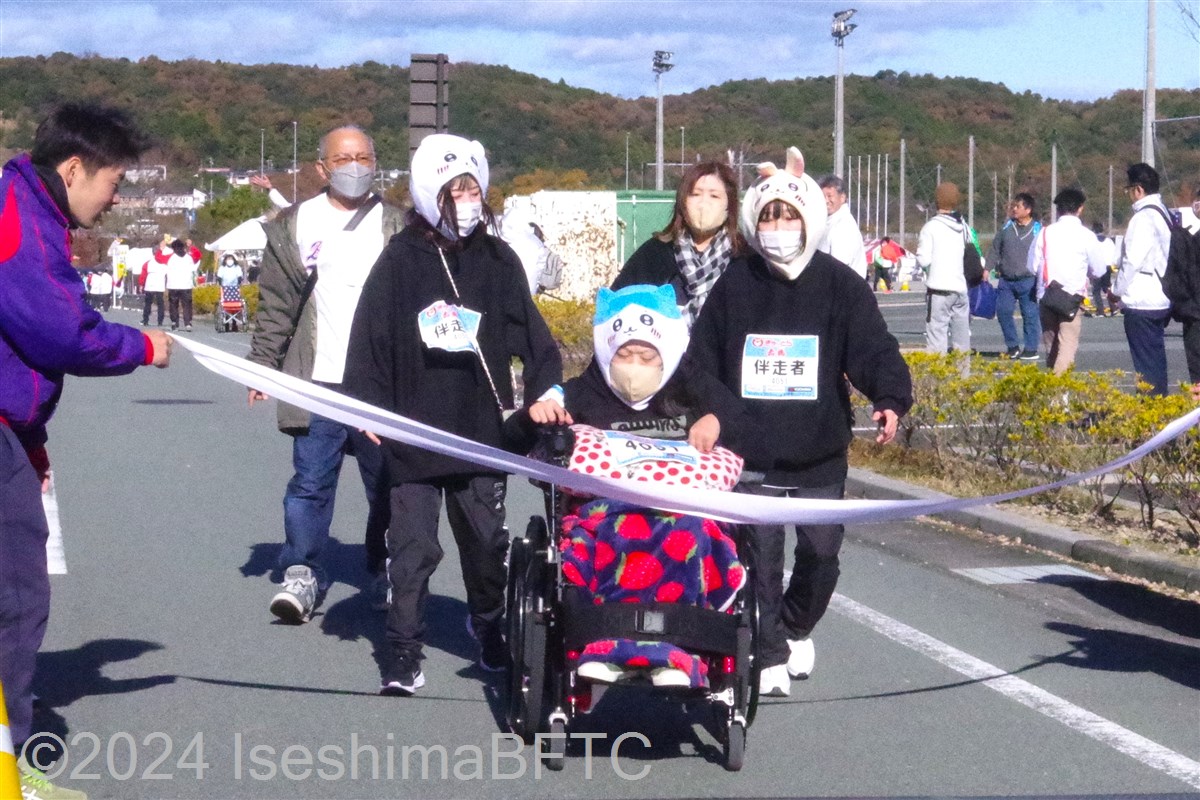 伴走者二人とともにかぶりものの仮装でゴールテープを切る車いす選手