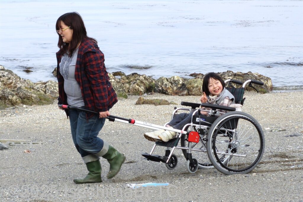 砂浜を、お母さんに「ジンリキ」を引いてもらって散歩する車いすの女の子