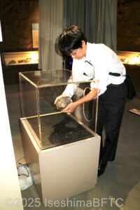 手が入れられるように丸くくりぬかれた透明の四角いケースの中の鉱石を男性が持ち上げているようす。
その重さを感じることができる。