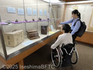 鈴のコレクションを眺める女性2名。1名は車いすに乗っている。