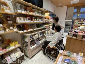 壁一面に陳列された商品。膝に買い物カゴを乗せた車いすの女性が、高いところにある商品を、スタッフに取ってもらっているようす。