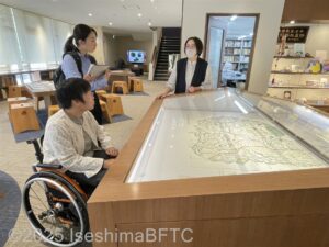 展示用ケースに入った地図を前に、学芸員が女性2名に説明をしているようす。女性のうち一人は車いすに乗っている。