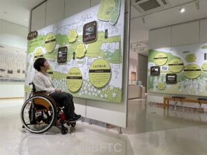 壁一面に展示されているパネルを見ている車いすの女性のようす。
