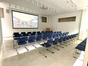 松阪ものがたりシアターと呼ばれる視聴覚コーナー。大きなスクリーンの前に、青色のイスが30脚ぐらい置かれている。