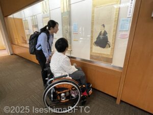 本居宣長の自画像が描かれている掛け軸を眺める女性2名。1名は車いすに乗っている。