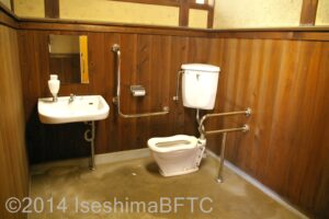 車いす対応トイレの室内のようす。便器に向かって左手側奥の壁にはL字手すりが付いていて、右側にはU字の手すりが付いている。