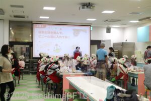 「ホシオくんキッチン」での体験の説明を受ける子供たち。子供たちは赤いエプロンとカラフルな帽子を被っている。