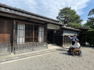 古い日本家屋の旧宅の前で並ぶ女性2名。一人は車いすに乗っている。