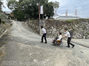 資料館に続く上り坂を男性二人がじんりき/けん引式車いす補助装置を使って車いすを介助しているようす。
