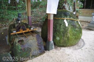 手水舎のようす。手水舎の桶は石でてきている。そのすぐ横に直径1mほどの苔むした丸い石が置いてあり、しめ縄が巻かれている。