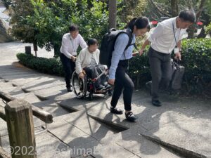 駐車場から急な坂道を、JINRIKI（じんりき/けん引式車いす補助装置）を使って3名で介助しているようす。