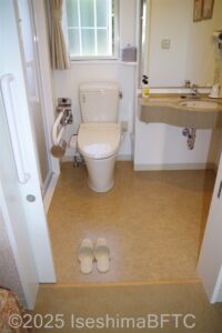 部屋の車いす対応トイレ入り口からのようす　正面に便器があり左がわにU字の手すり　右側には洗面