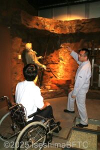 当時の手彫り採掘を再現した展示物を前に、資料館のスタッフが車いすの男性に説明をしているようす。