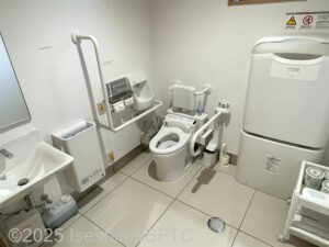 車いす対応トイレの室内のようす。便器に向かって左側の壁にはL字手すりが付いていて、右側にはU字の可動式手すりが付いている。ベビーシートもあり。