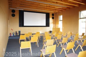 展示棟内にある映像ホールのようす。正面に大きなスクリーン。その前に木製のイスが30脚ぐらい置かれている。