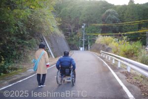 国道から江の浦トンネル北側入口へまで道を歩く男女。男性は車いすに乗っている