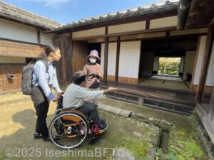 公開住居　後庭から室内を眺めている女性二人。一人は車いすに乗っている。横に立つボランティアが説明をしているようす。