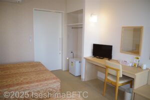 室内のベッド側から部屋の全景　机の上にテレビその左横には正方形の小さな冷蔵庫　奥には浴室トイレのドア