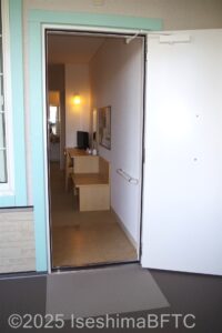 バリアフリールームの入り口　入り口入ってすぐ右の壁に室内に続く手すり　その先にテレビのある棚と机