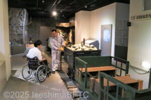 展示物を前に、資料館のスタッフが車いすの男性に説明をしているようす。