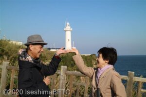 公園から灯台方面を見たようす。60代の視覚障害者のカップルが、向かい合って手を前に出し、手のひらを上に重ね合い、その上に灯台が乗っているように見える写真。