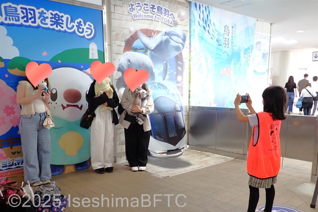 ぬいぐるみを持ってポーズを取る女性3名をスマホで撮影している小学生ボランティア