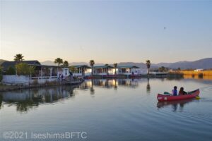 夕景の湖に赤いカヌーが浮かんでいる。その向こう側にグランピングのキャビンが見える。