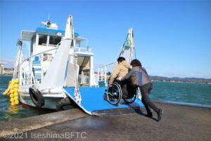 桟橋から福寿荘専用の船に設置されているスロープで乗船する車いすの男性と介助する女性のようす。