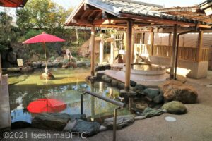 庭園露天風呂のようす。岩風呂と、木製の円形浴槽がある。周りは日本庭園のようなつくりで、赤いのだて傘が見える。