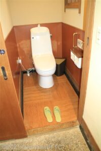 脱衣所内にある洋式トイレのようす。短いグリップ手すりあり。温水洗浄便座付（操作パネル：便座横）。