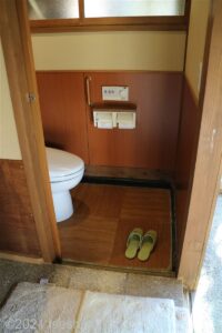 脱衣所内トイレ入口片開きドア。短いグリップ手すりあり。温水洗浄便座付（操作パネル：便座横）。