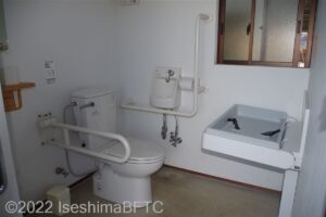 車いす対応トイレの室内のようす。便器に向かって右側奥の壁にはL字手すりが付いていて、左側にはU字の可動式手すりが付いている。ベビーシートも設置されている。