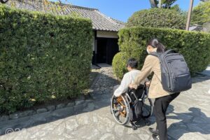 車いすの女性が介助してもらいながら、生垣の間の通路を通って住居へ向かおうとしているようす