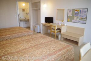 ベッド側からの部屋の全景　ベッドの向かい側にデスク、その左横にクローゼットと小さな冷蔵庫