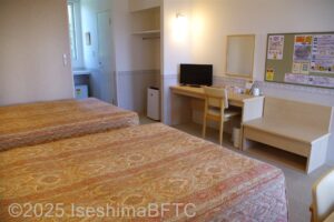 ベッド側からの部屋の全景　ベッドの向かい側にデスク、その左横にクローゼットと小さな冷蔵庫