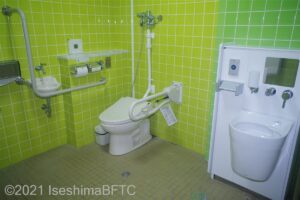 トイレ室内のようす　右にオストメイト　左に便器　便器に座ると右側L字手すり　左側U字可動手すり　右壁にトイレットペーパーホルダー　その上の棚壁に温水洗浄便座操作パネルがある