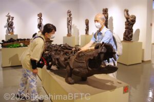 ワニのような大きな彫刻にさわる、白杖を持った視覚障害者