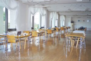 レストランのようす。ダイニング用のイスとテーブルがたくさん並んでいる。窓には白いカーテンがかかっている。