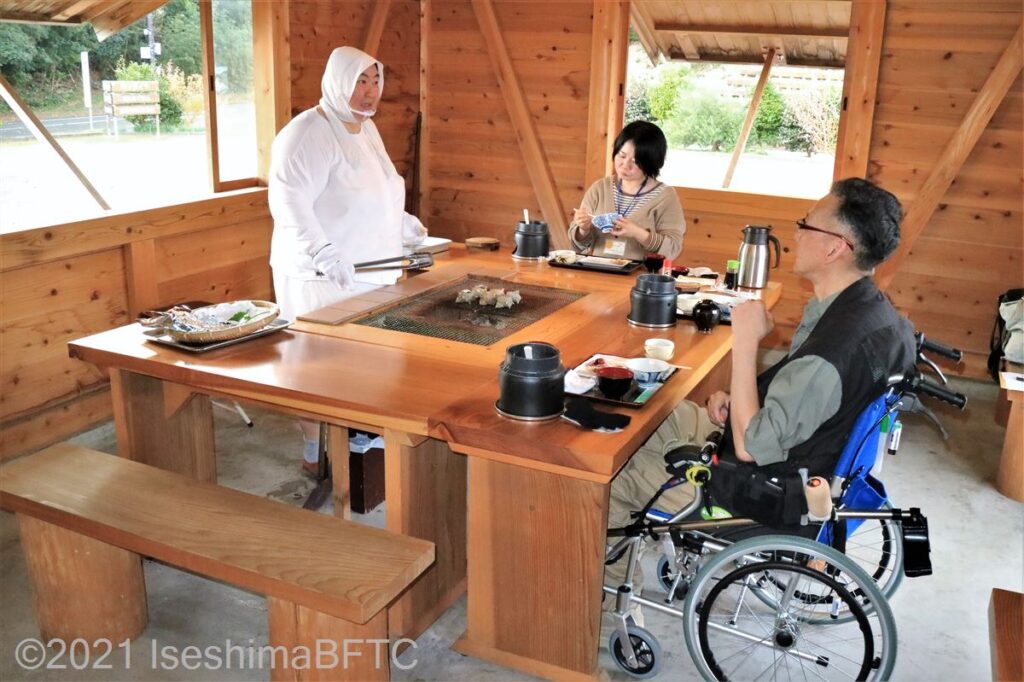 さとうみ庵での食事風景。男性と女性がテーブルにつき、白い磯着を着た海女さんが給仕をしているようす。男性は車いすに乗っている。