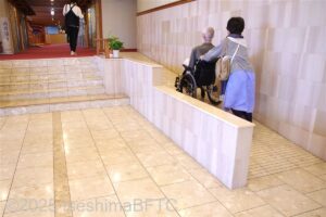エレベーター方面へのスロープを介助してもらって上がる車いすのお客さま