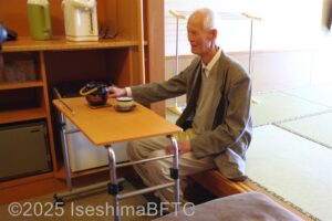 413号室　ベッド用テーブルに湯呑みを置き、和室の小上がりに腰掛けてお茶を飲むお客さま
