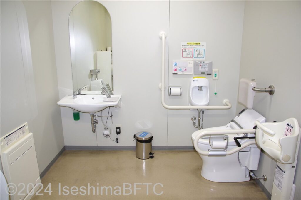 志摩市役所1階車いす対応トイレ　入口からみて正面奥に、よこ置きに便器。入口からみて左奥に洗面台