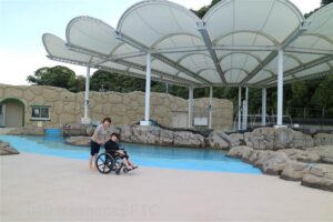 浜に見立てた人口のプール。上には大っきな白い屋根がかかっている。その前でこちらを向く女性2名。右側の女性は車いすに乗っている。